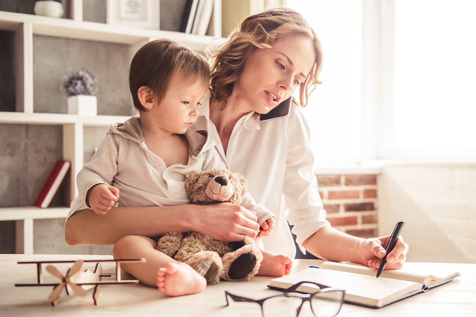 A woman, multitasking her mommy makeover, holds a toddler cuddling a teddy bear while talking on the phone and jotting notes in a notebook at her desk. The room is lined with bookshelves, and a small model airplane sits on the desk. Her glasses are close at hand.