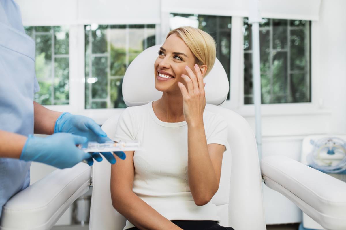 A woman with blonde hair sits on a white chair in a clinic, smiling at a healthcare provider wearing gloves and scrubs, who is holding a medical tray. Large windows with greenery outside hint at the serene setting of her plastic surgery consultation.