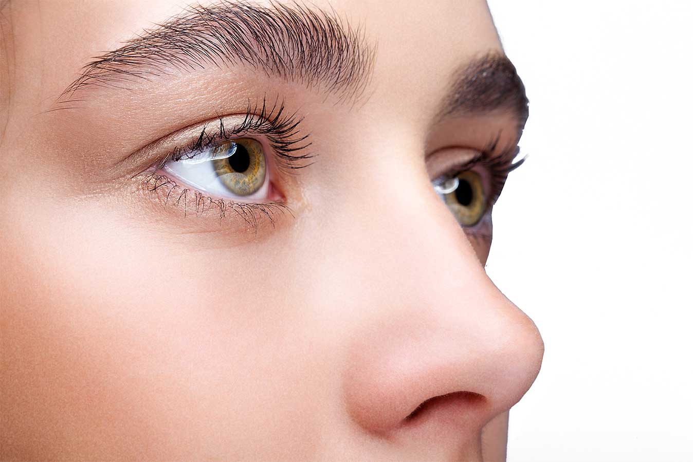 Gros plan sur le visage d'une personne, mettant l'accent sur le nez et les yeux, révélant une peau lisse et des yeux marron clair. L'image, potentiellement post-rhinoplastie, souligne les détails des cils et des sourcils sur un fond blanc uni.