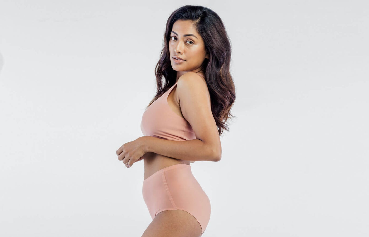 In a neutral-toned two-piece outfit, the woman exudes confidence as she poses against a plain background. With her long, wavy hair flowing and a slight smile, she embodies the elegance of a mommy makeover in this minimalistic setting.