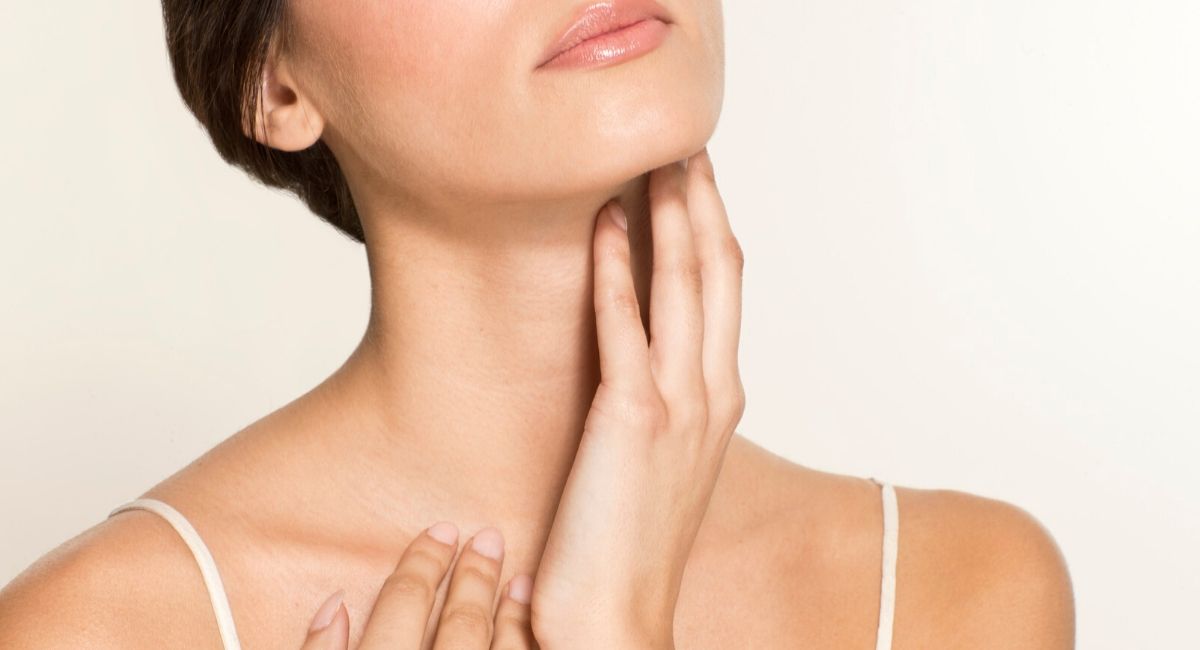 Close-up of a woman gently touching her neck and collarbone, with the other hand resting on her chest. She showcases smooth skin, free of submental fat, while wearing a white spaghetti strap top. The background is light and blurred.
