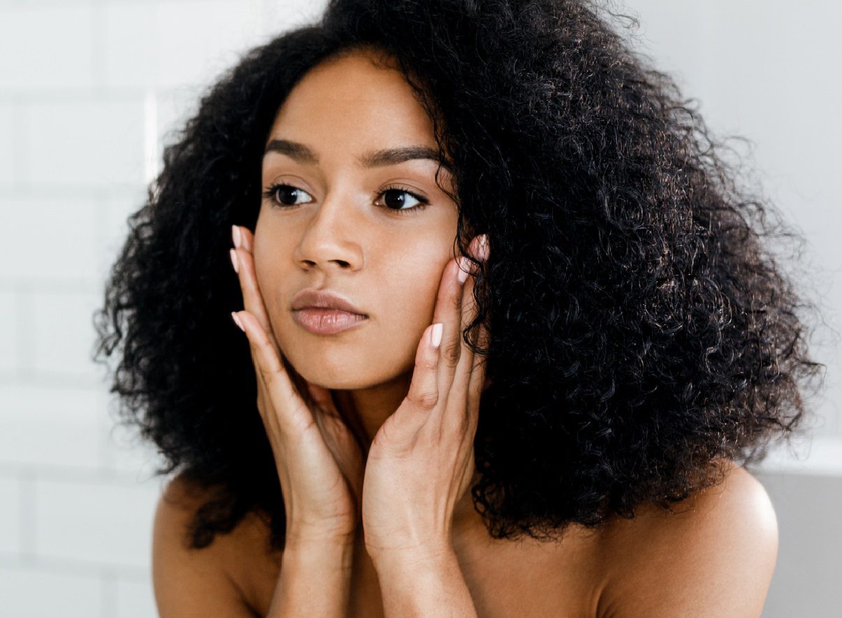 A person with curly hair gently holds their face with both hands, appearing thoughtful, perhaps contemplating the benefits of microdermabrasion. They have a neutral expression and look slightly away. The softly blurred background draws attention to their face.
