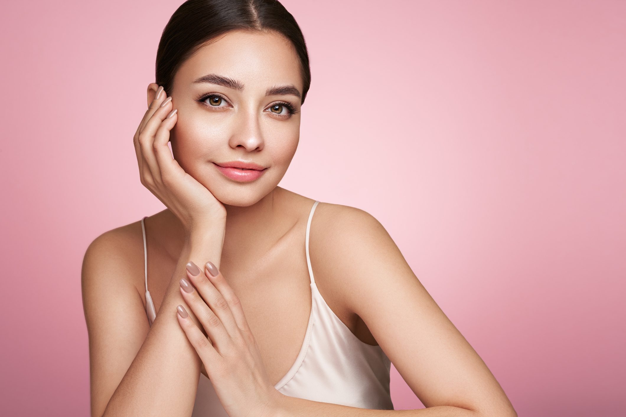 Une personne aux cheveux foncés pose, une main sur la joue, avec un doux sourire. Elle porte un débardeur clair sur un fond rose. Ses ongles sont manucurés et le soin du dermaplaning donne à sa peau un aspect particulièrement lisse et éclatant.