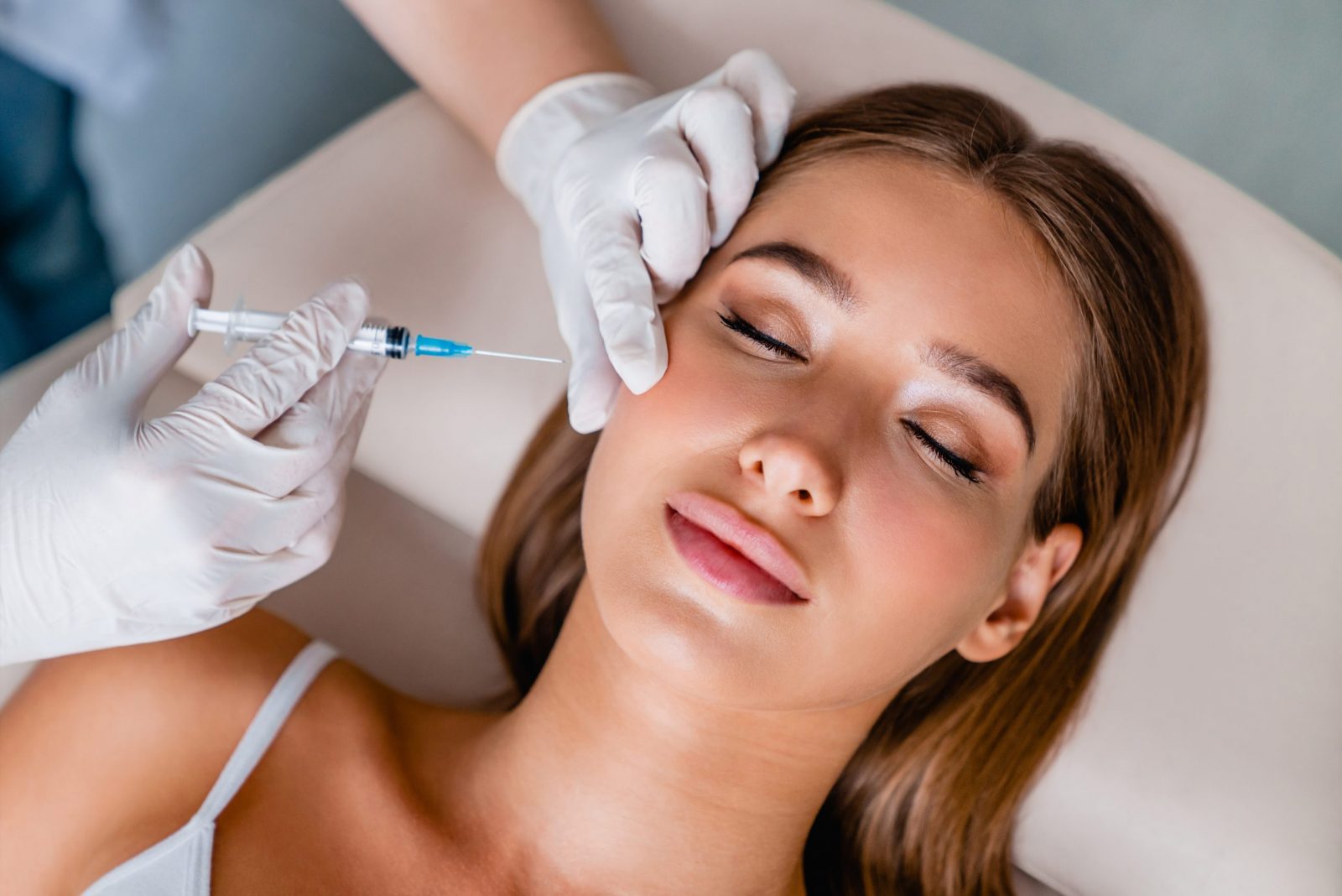 Person with closed eyes receiving a facial injection. A gloved hand gently supports their head while another administers the shot, performing a soft lift. The person lies back on a surface, looking relaxed and peaceful.