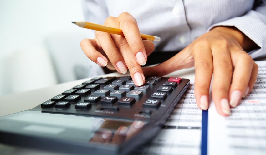 Close-up of a person using a calculator with one hand while holding a pencil in the other hand. The background shows printed documents, indicating a financial or budgeting task.