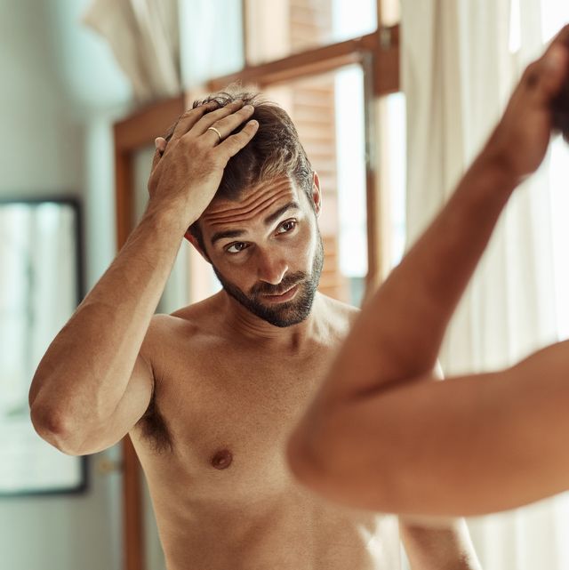 A shirtless man with short hair and a beard examines his reflection in a mirror, contemplating hair loss treatment. Indoors, he gently touches his hair with one hand. The room features large windows with sheer curtains, allowing natural light to illuminate the scene.
