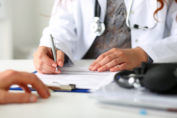 A doctor sits at a desk, writing on a clipboard with a pen. A stethoscope hangs around their neck while medical documents and a blood pressure monitor lie nearby. Another persons hand rests on the desk, perhaps inquiring about the intricacies and options surrounding plastic surgery costs.