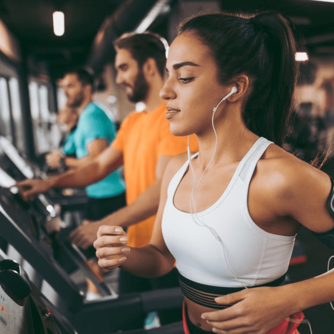 A woman in a white sports bra and earphones runs on a treadmill, focused and determined. In the well-lit gym, where natural light streams through large windows, two men and another woman exercise nearby, each possibly inspired by their personal fitness goals rather than liposuction.