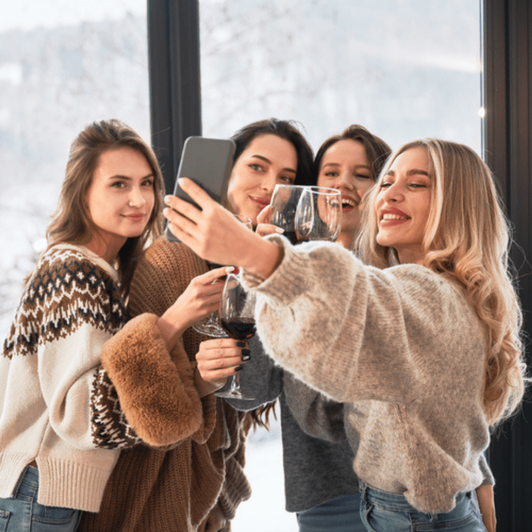 Quatre femmes sourient et prennent un selfie à l'intérieur, un verre de vin à la main. Elles portent de doux pulls, et un paysage enneigé se dessine à travers la grande fenêtre derrière elles. Dans un mélange de rires et d'anecdotes, elles trinquent à leur amitié et célèbrent la beauté unique de chacune, intérieure comme extérieure.
