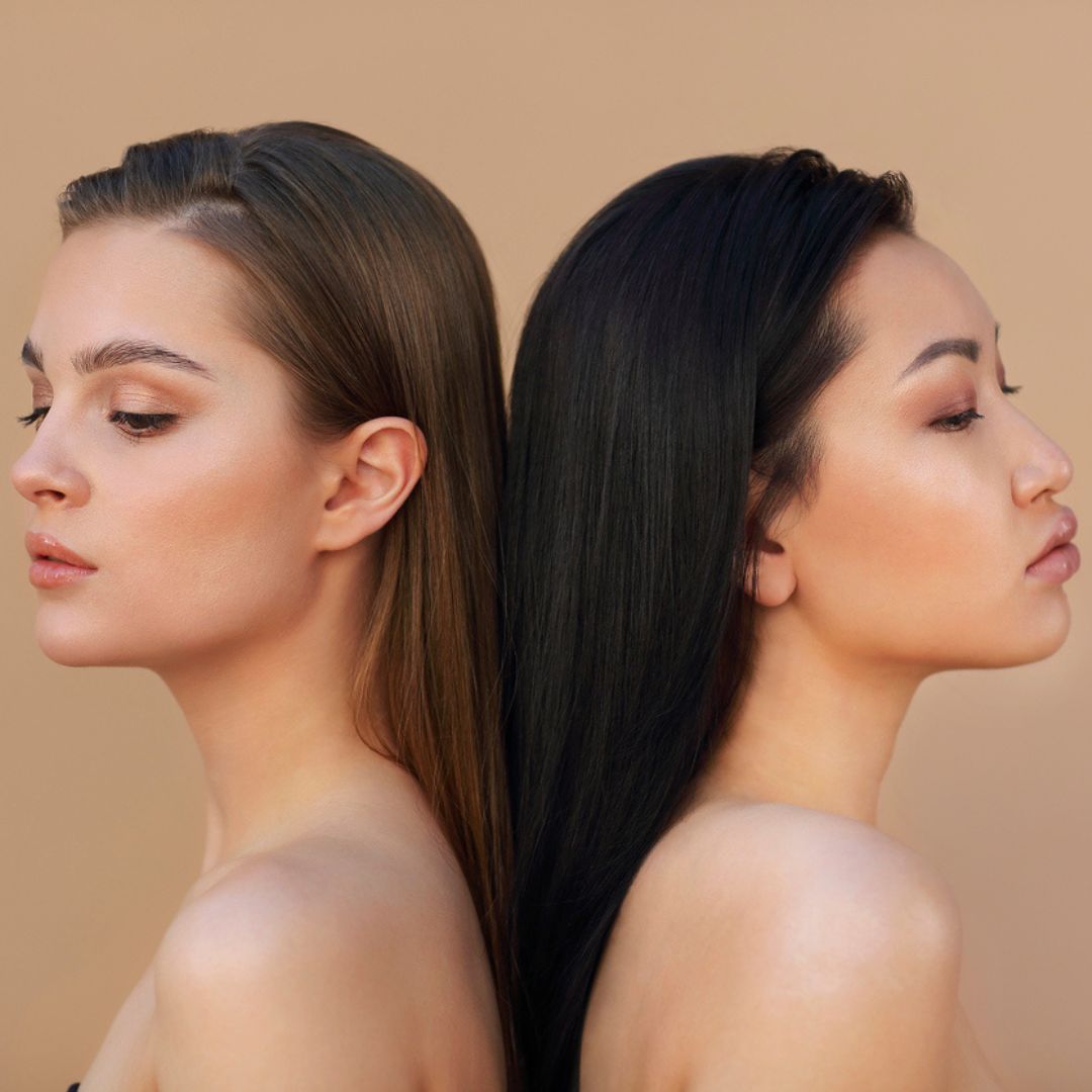 Two women with smooth, straight hair stand back-to-back against a beige background, showcasing their refined profiles. The woman on the left has light brown hair, while the one on the right boasts black tresses. Their serene expressions hint at elegance and confidence, reminiscent of buccal fat removals transformative touch.