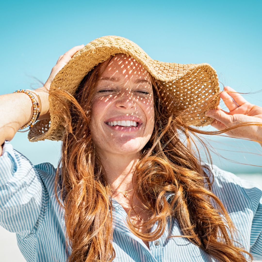 Smiling woman with long red hair wearing a sunhat and a striped shirt, standing on a sunny beach. The hat casts a pattern of light and shadow across her face, subtly concealing signs of rosacea. Blue sky and ocean are visible in the background.