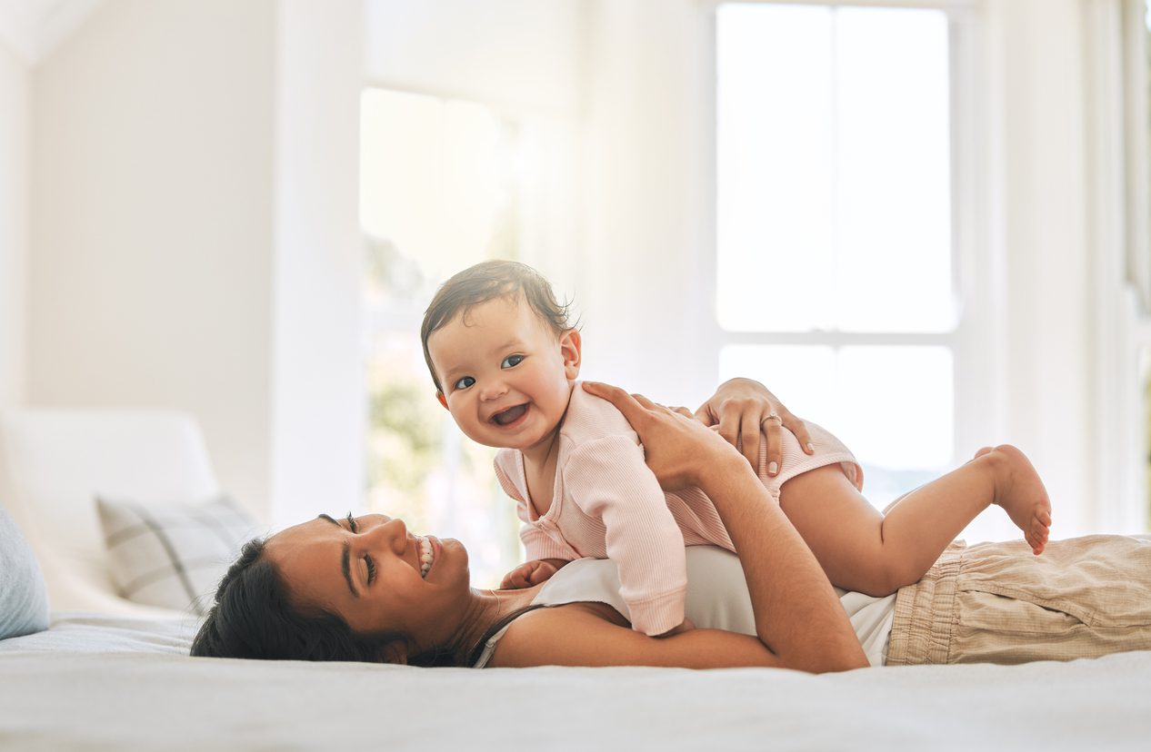 A woman lies on her bed, smiling up at a baby who happily rests on her stomach, which shows the immaculate results of abdominoplasty. The room is bright with natural light streaming through the windows in the background.