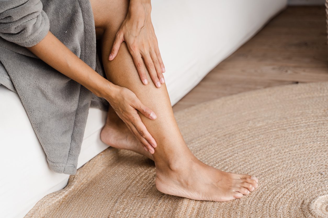 A person sits on a white couch, gently massaging their bare leg affected by varicose veins with both hands. They are wearing a gray robe, and the floor is covered with a textured beige rug. The setting appears calm and relaxed.