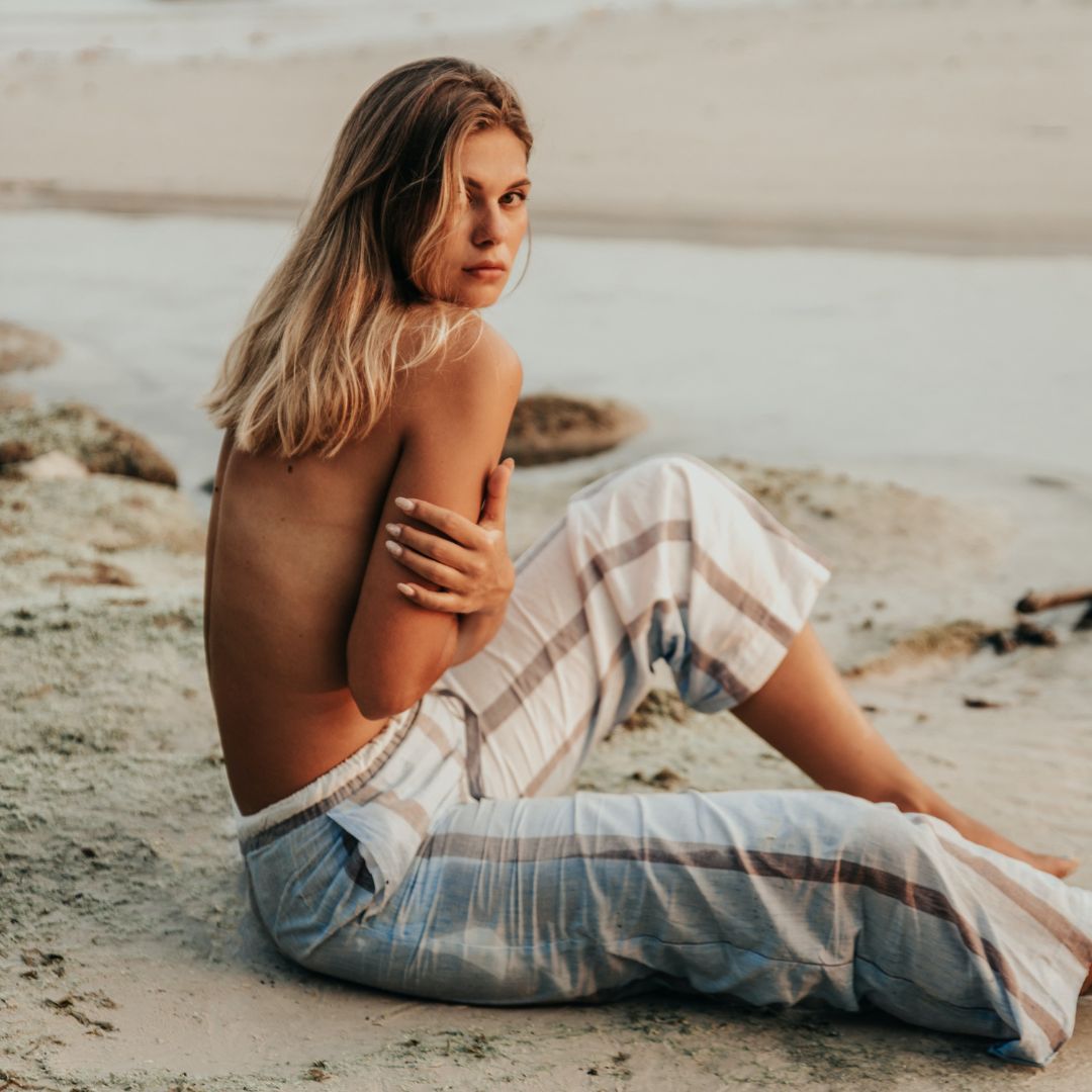 A woman with long blonde hair sits on a sandy beach, turned slightly towards the camera. Shes wearing striped pants and has her arms crossed over her chest, exuding serenity and confidence. The beach and ocean provide a soothing backdrop as she reflects on her recent journey of nipple correction surgery.