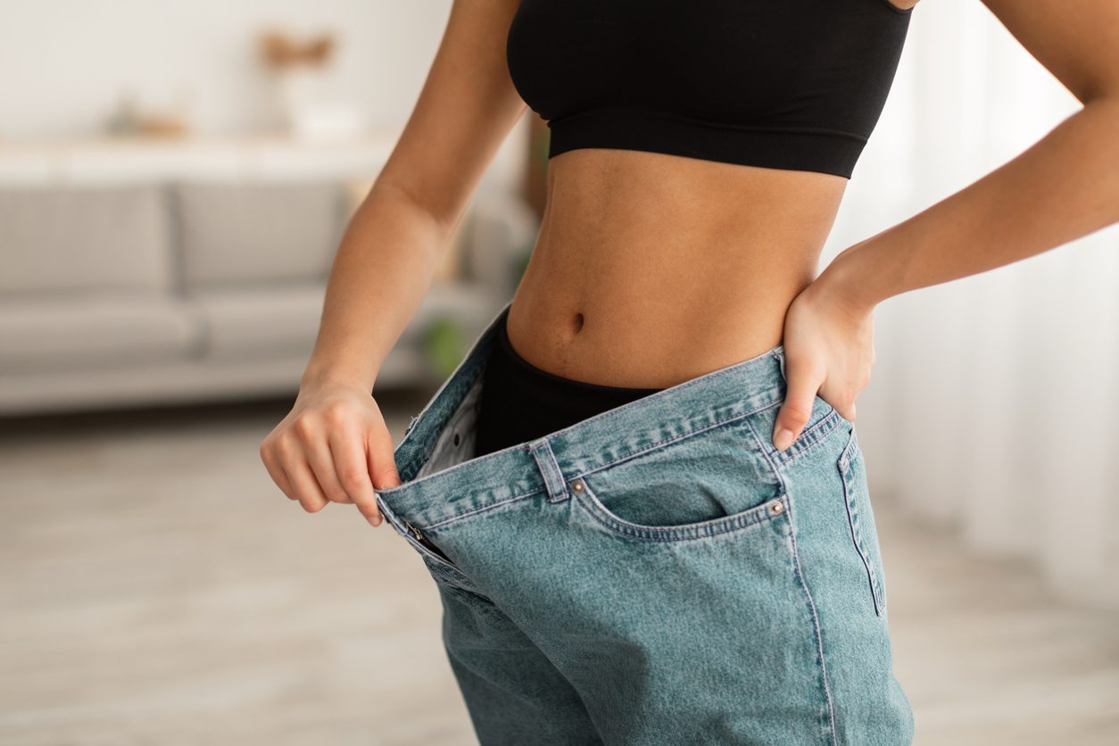 A person in a black crop top highlights their weight loss by pulling the waistband of loose jeans, possibly after a gastric sleeve procedure. The rooms blurred background reveals a sofa and light curtains.