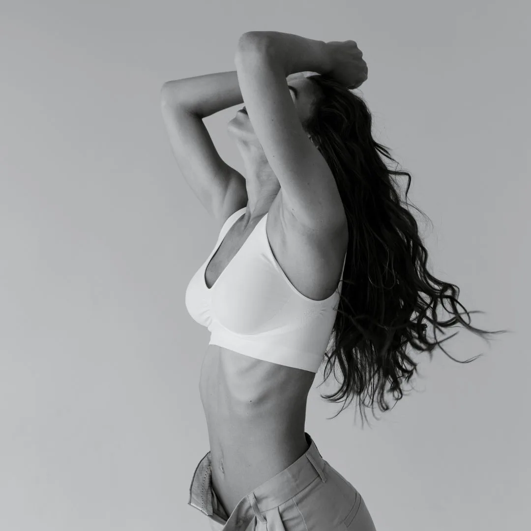 A woman with long wavy hair, showcasing her confidence in a white sports bra and light-colored pants, poses against a plain background. Her arms are raised above her head, enhancing the elegance of her form as she gazes upward. The image, captured in black and white, subtly highlights natural beauty and poise.
