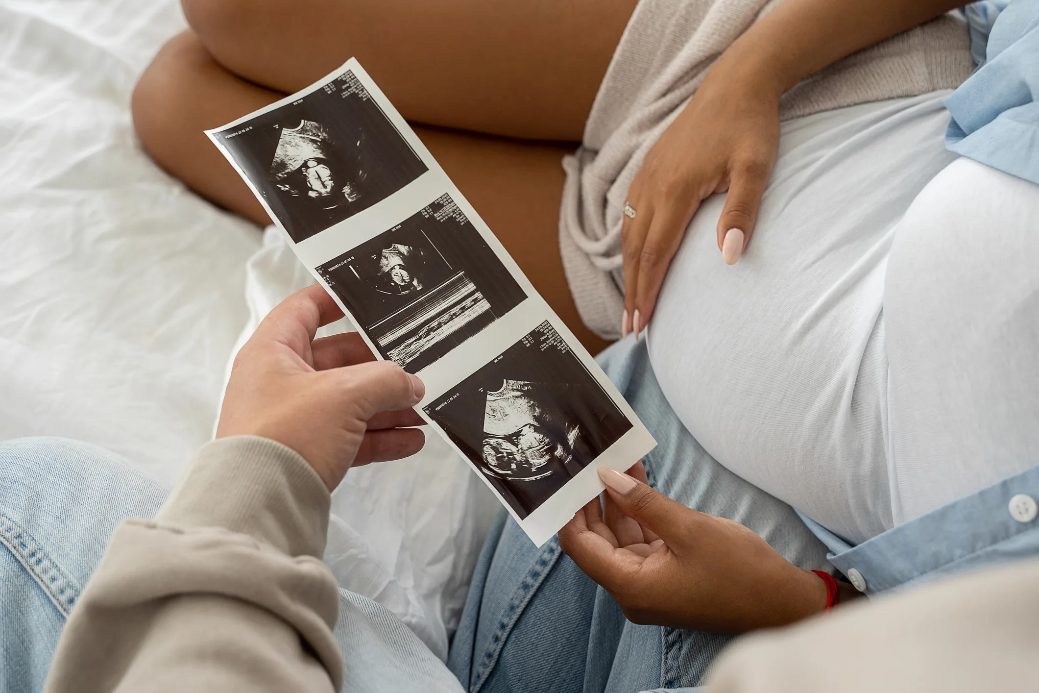 Two people on a bed holding ultrasound images. One person, with hand on their belly, wears a white top and beige cardigan. The other, in light jeans and a sweatshirt, holds the image strip. The scene suggests an expecting couple reviewing the fruits of their In Vitro Fertilization journey.