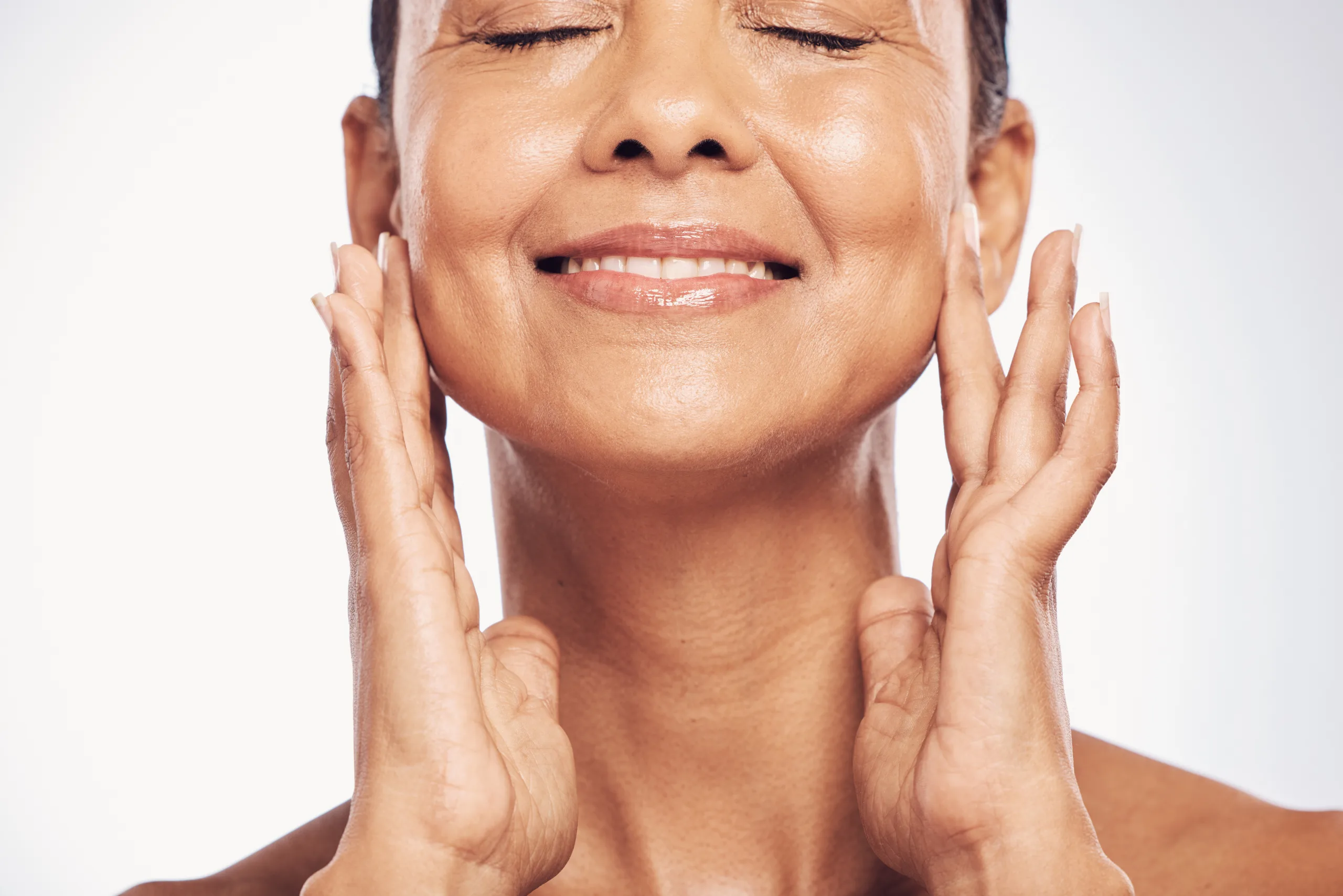 A person with closed eyes smiles and gently touches their face, revealing skin made smooth and radiant through Thermage® treatments. The background is a soft, light blue, creating a serene atmosphere.