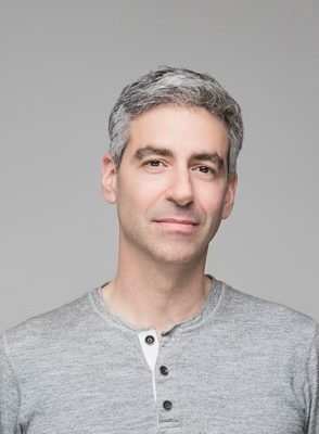 A person with short gray hair wearing a gray henley shirt, standing against a plain light gray background, appears to have just experienced the rejuvenating effects of phenol peels. The image focuses on their upper body and face, highlighting a fresh and radiant complexion.