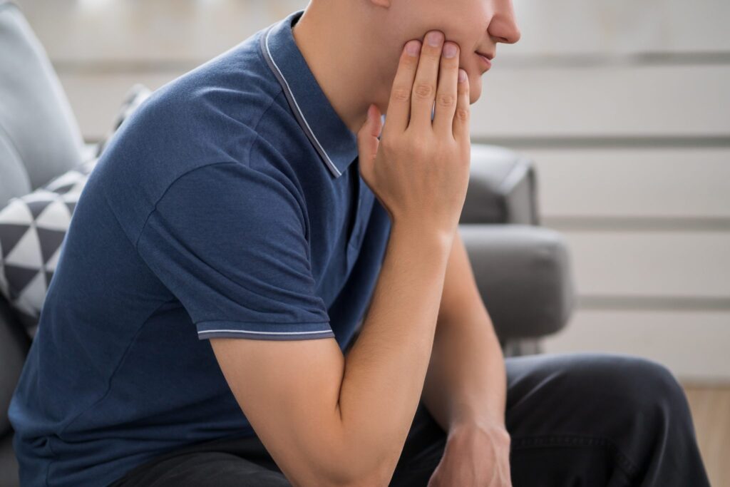 A person in a blue polo shirt is sitting on a couch, holding their cheek with a pained expression, possibly experiencing discomfort from recent wisdom teeth removal. A pillow with a geometric pattern is visible in the background.