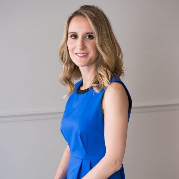 A woman with long blonde hair, wearing a blue sleeveless dress, poses gracefully against a light grey background. She is smiling and looking towards the camera, radiating confidence in her decision to embrace change after undergoing labiaplasty.