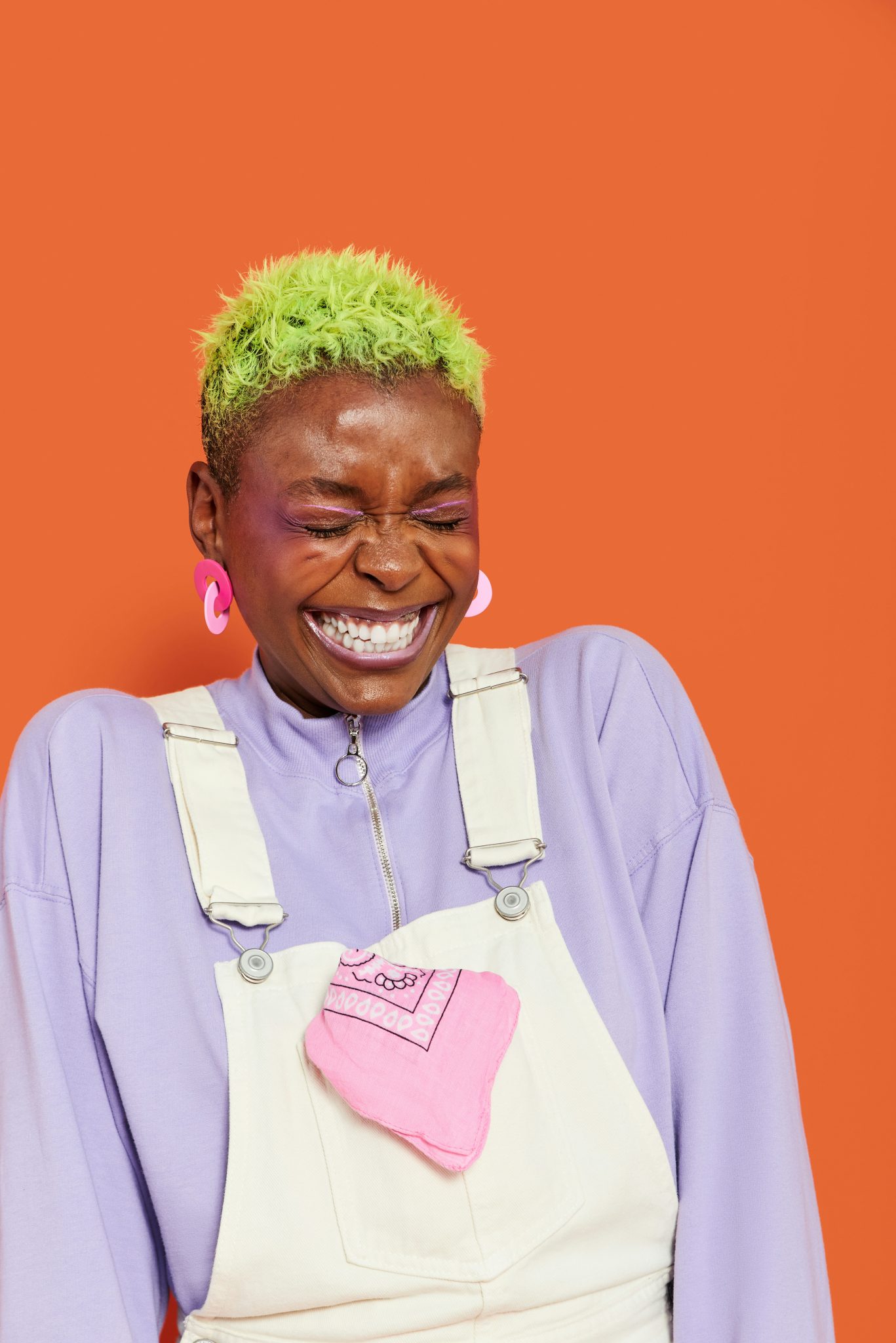 Person with vibrant green hair and pink earrings, wearing a purple shirt and white overalls with a pink pocket, is smiling broadly—thanks to cosmetic dentistry financing—with eyes closed against an orange background.