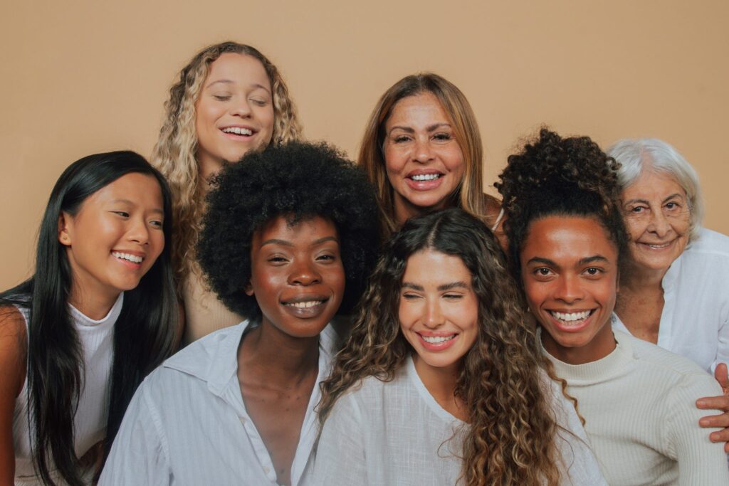 A diverse group of seven smiling women, representing data from the 2023 Procedural Statistics Report, includes older and younger individuals with various hair types and skin tones. Theyre posing in front of a beige background, wearing casual white and light-colored clothing.