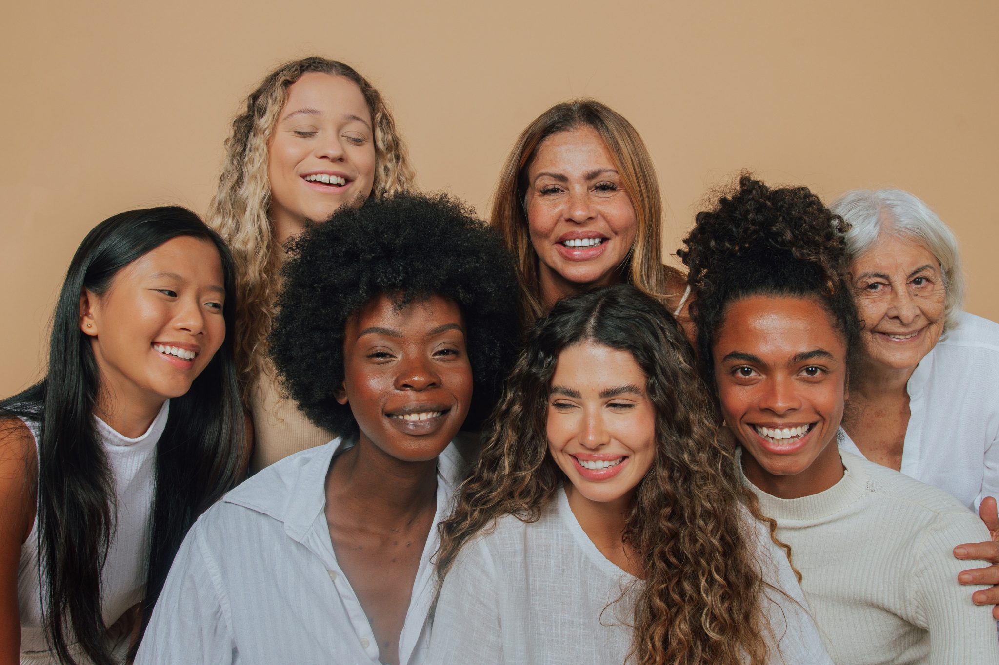 A diverse group of seven smiling women, representing data from the 2023 Procedural Statistics Report, includes older and younger individuals with various hair types and skin tones. Theyre posing in front of a beige background, wearing casual white and light-colored clothing.