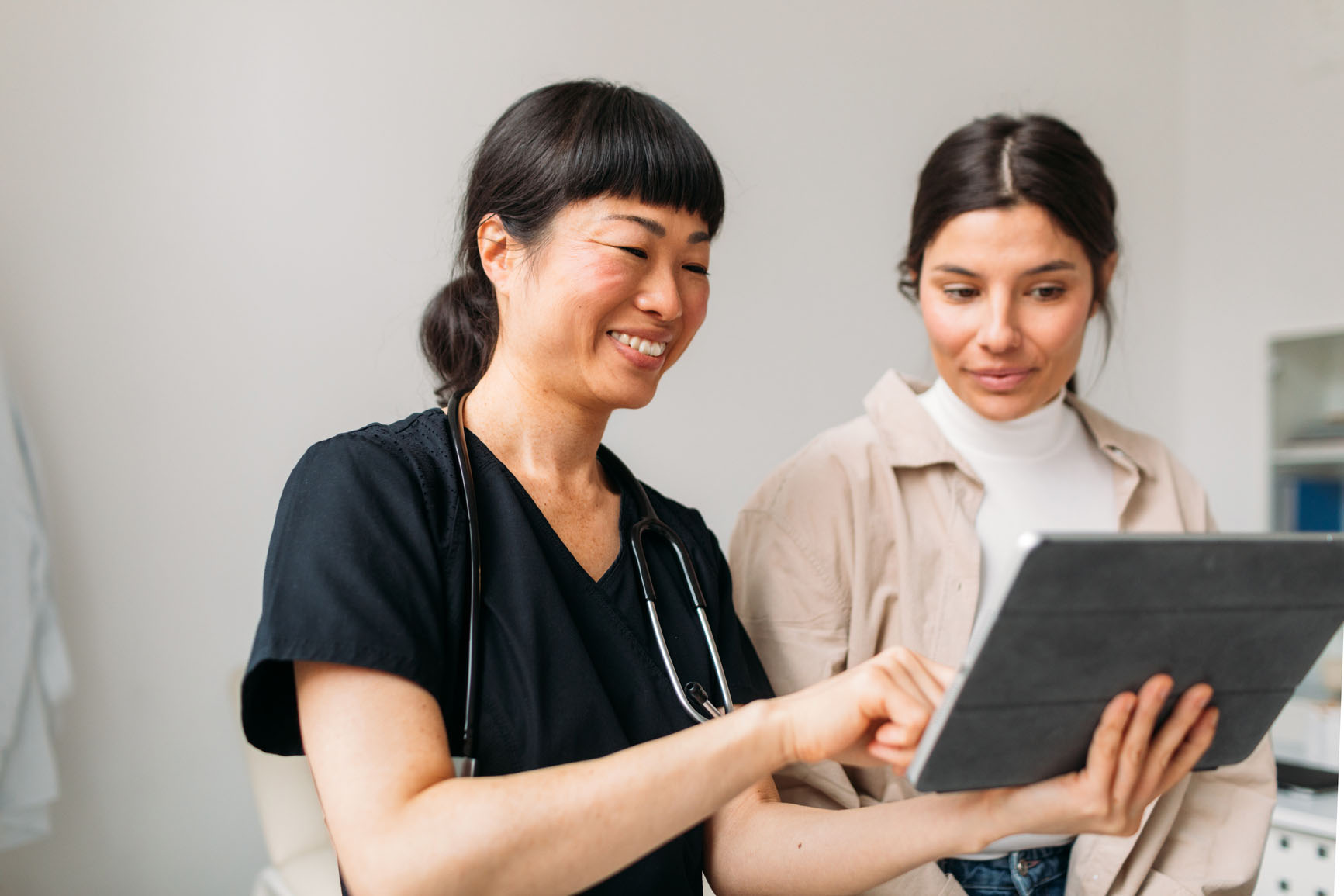 A healthcare professional in black scrubs shows information on a tablet to a woman in a beige jacket. They are smiling and standing in a bright, modern room. The healthcare professional has a stethoscope around their neck.