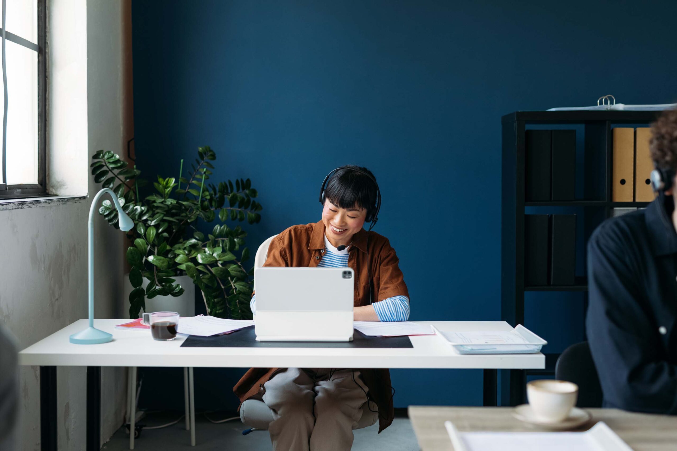 A person sits at a white desk using a laptop, wearing headphones and smiling. A plant and lamp are on the desk, with binders on a shelf against a blue wall in the background. A coffee cup is visible on a table in the foreground.