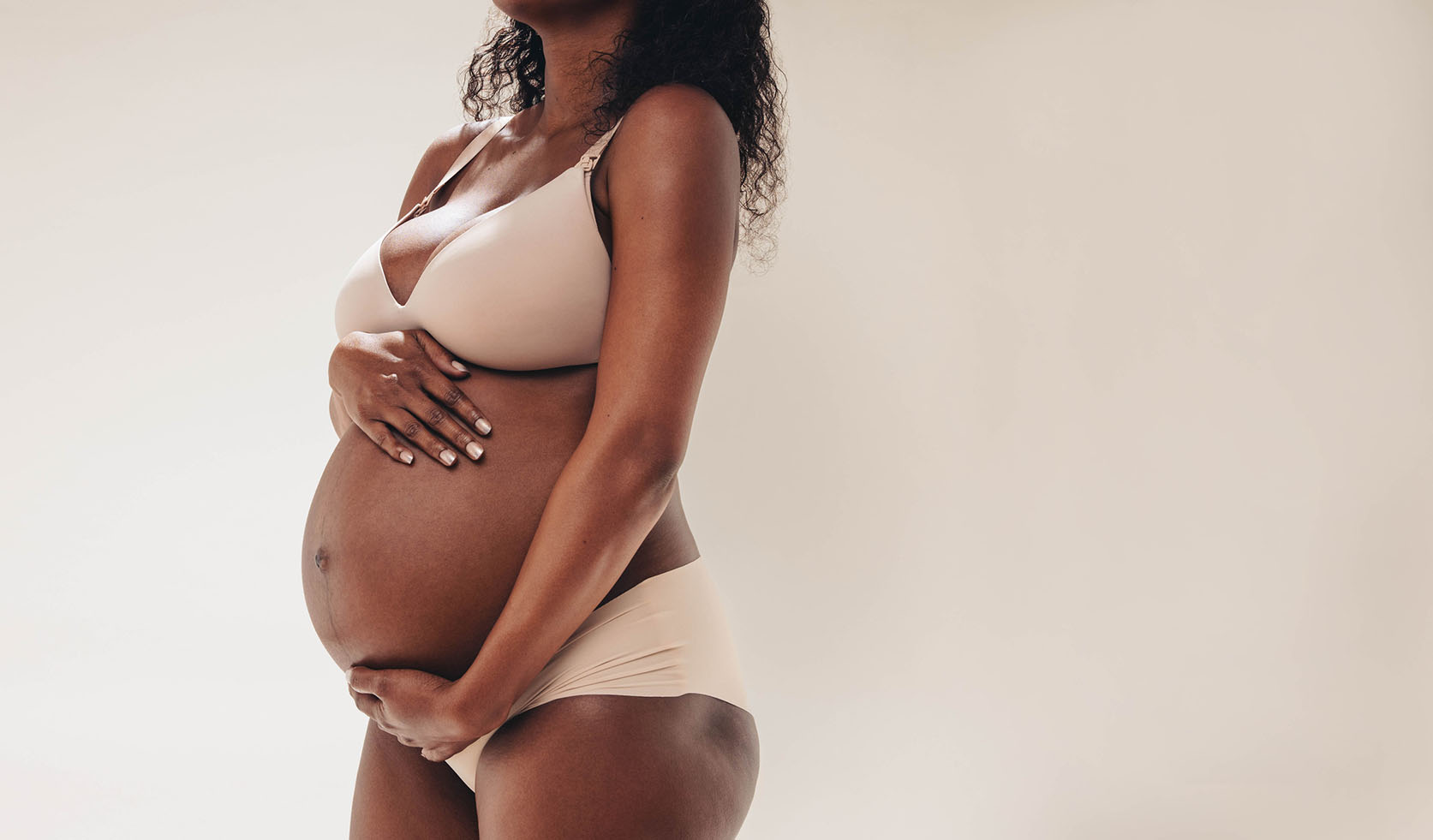 Pregnant woman in beige underwear cradling her belly with both hands, standing against a plain, light-colored background. Her face is not visible.