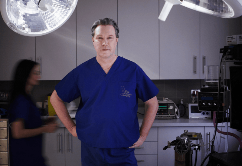A person in blue medical scrubs stands confidently in a surgical room, hands on hips. They are framed by bright surgical lights, highlighting the precision tools used for procedures like the Liquid Breast Lift™, with an indistinct figure blurred in the background.