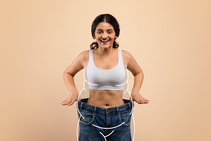 Smiling, fit woman celebrating weight loss success in yellow activewear on blue background.