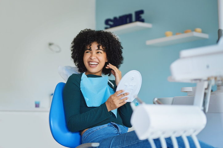 Smiling woman in a dental chair holding a mirror during an All on 4 dental implants consultation in a modern clinic.