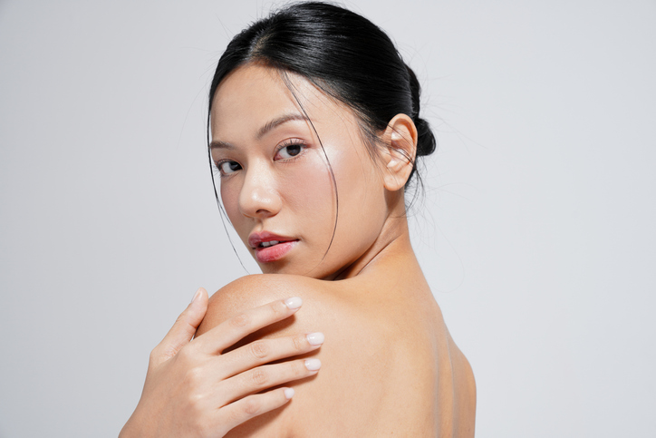 Young woman with glowing skin looks over her shoulder, hand resting on bare shoulder, in a minimal studio setting.