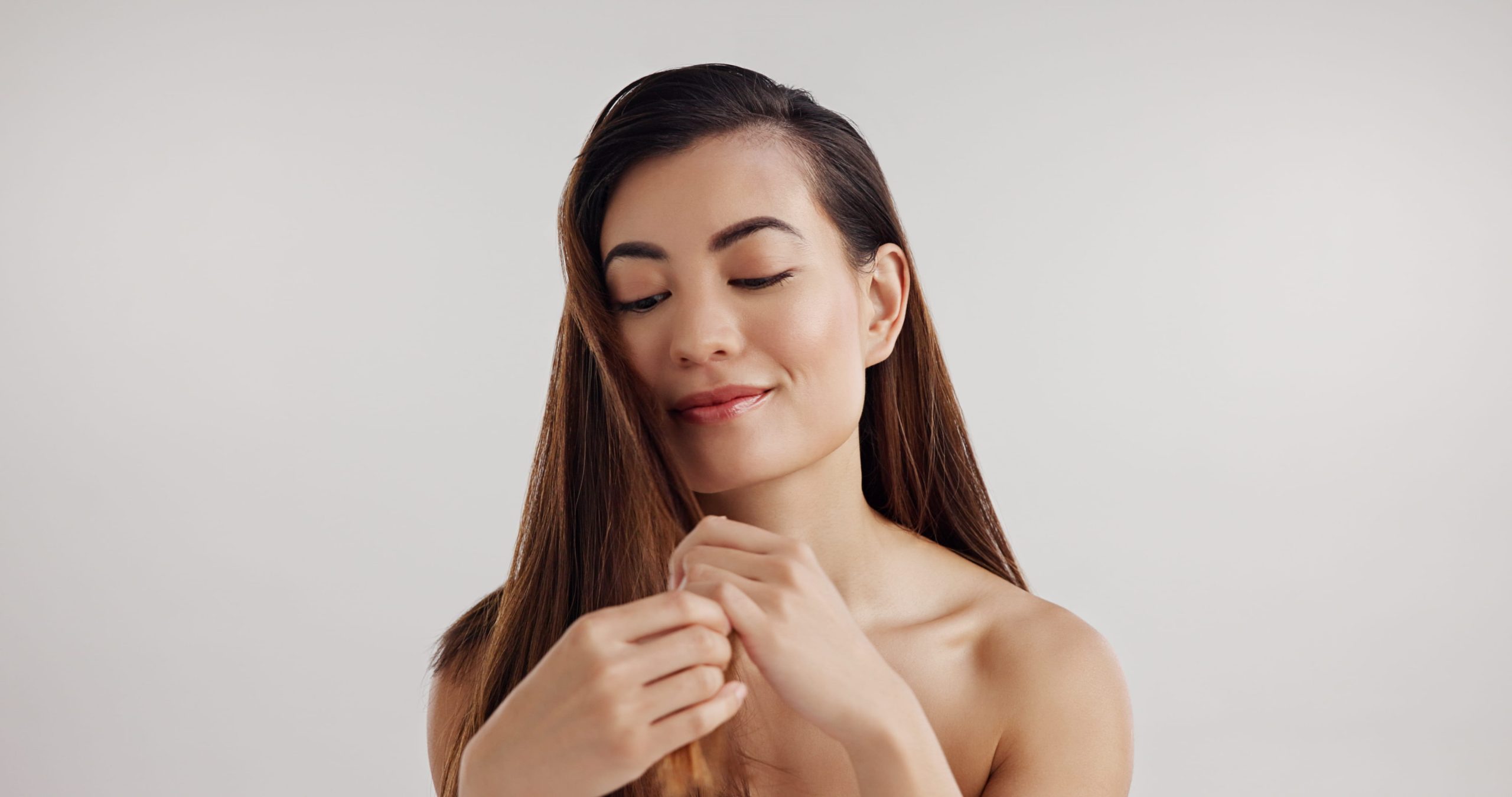 Woman with long straight brown hair gently holding a section of her hair against a neutral background.