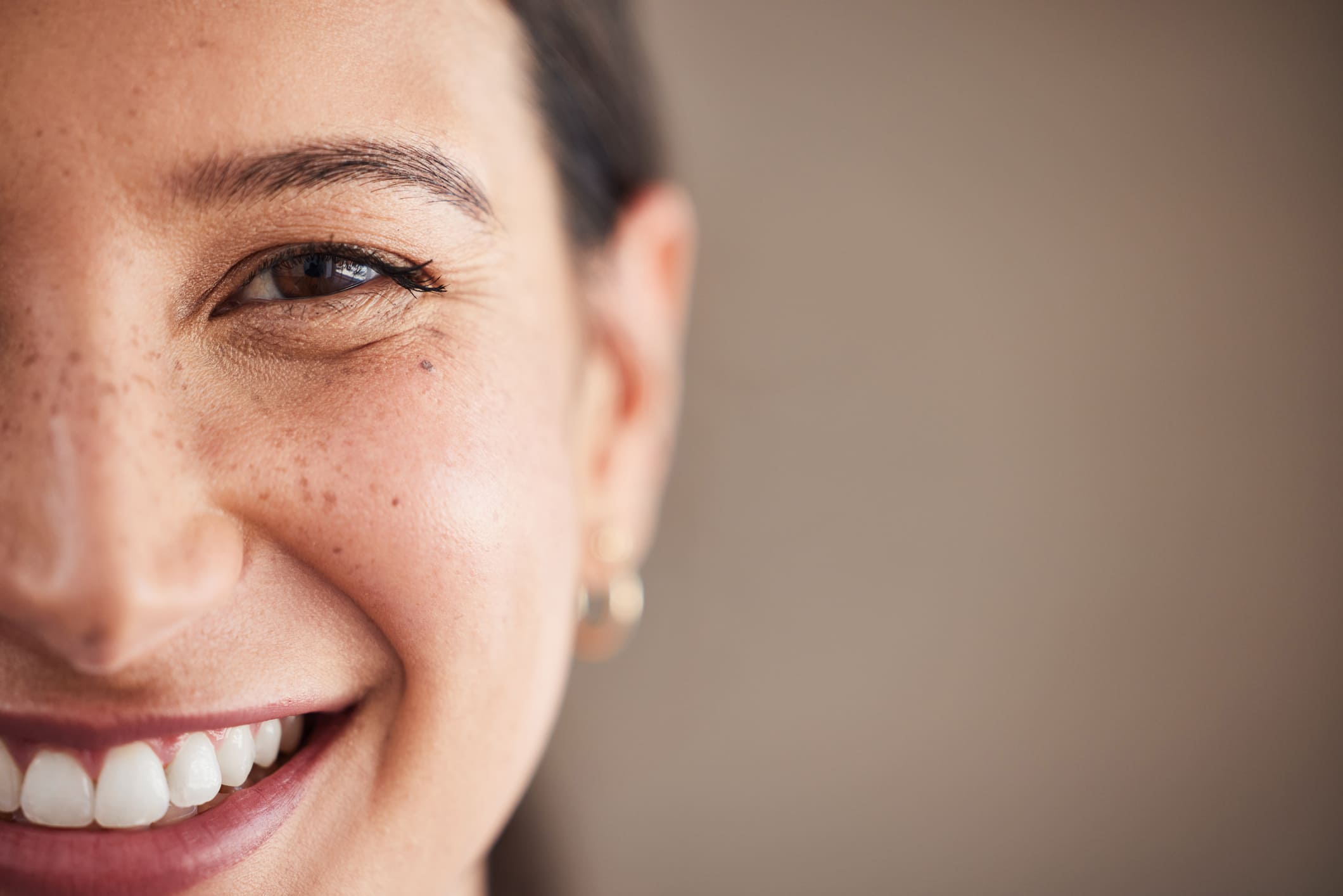 Close-up portrait of a smiling woman with natural skin, freckles, and visible teeth against a soft neutral background.